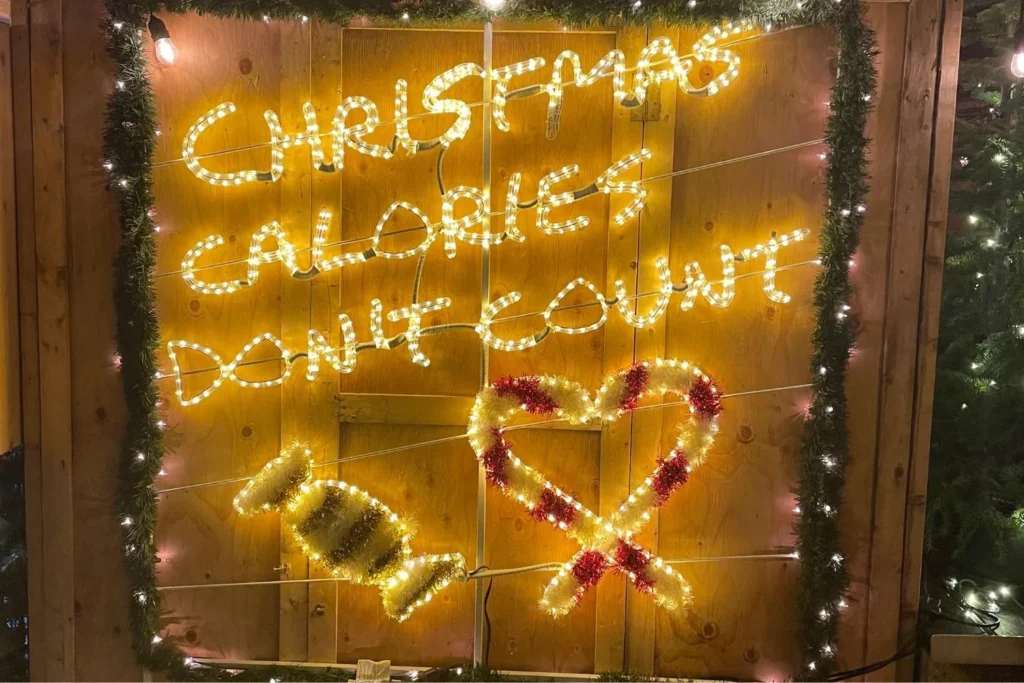 Bright festive sign made of string lights reading "Christmas Calories Don’t Count" with a candy and heart-shaped design, adding playful energy to the holiday dining atmosphere.