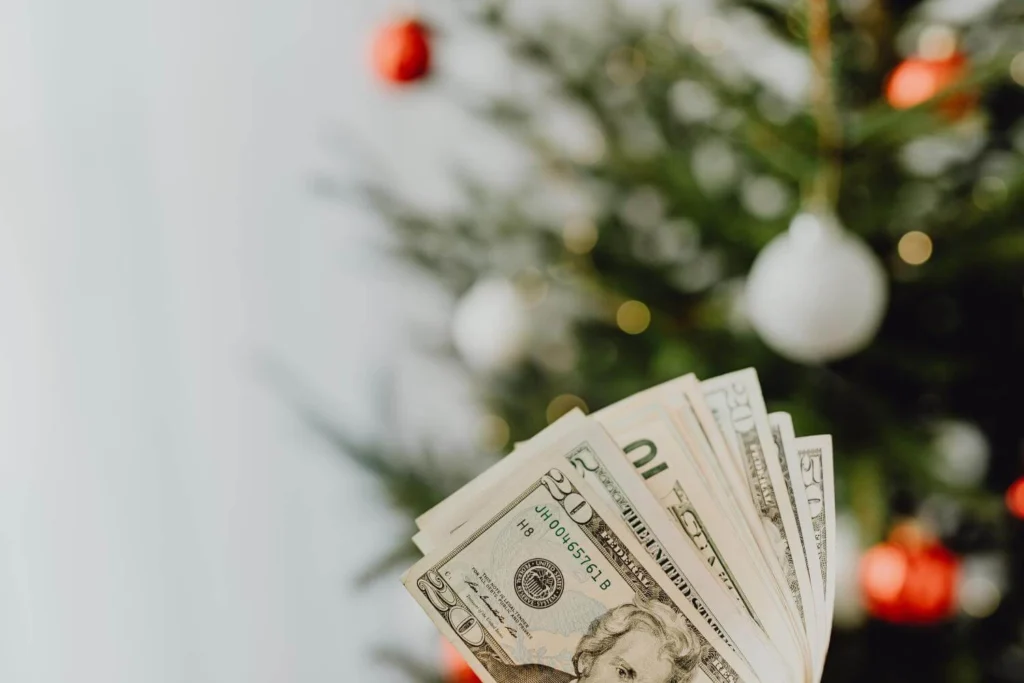 Fanned-out US dollar bills held in front of a decorated Christmas tree with red and white ornaments.