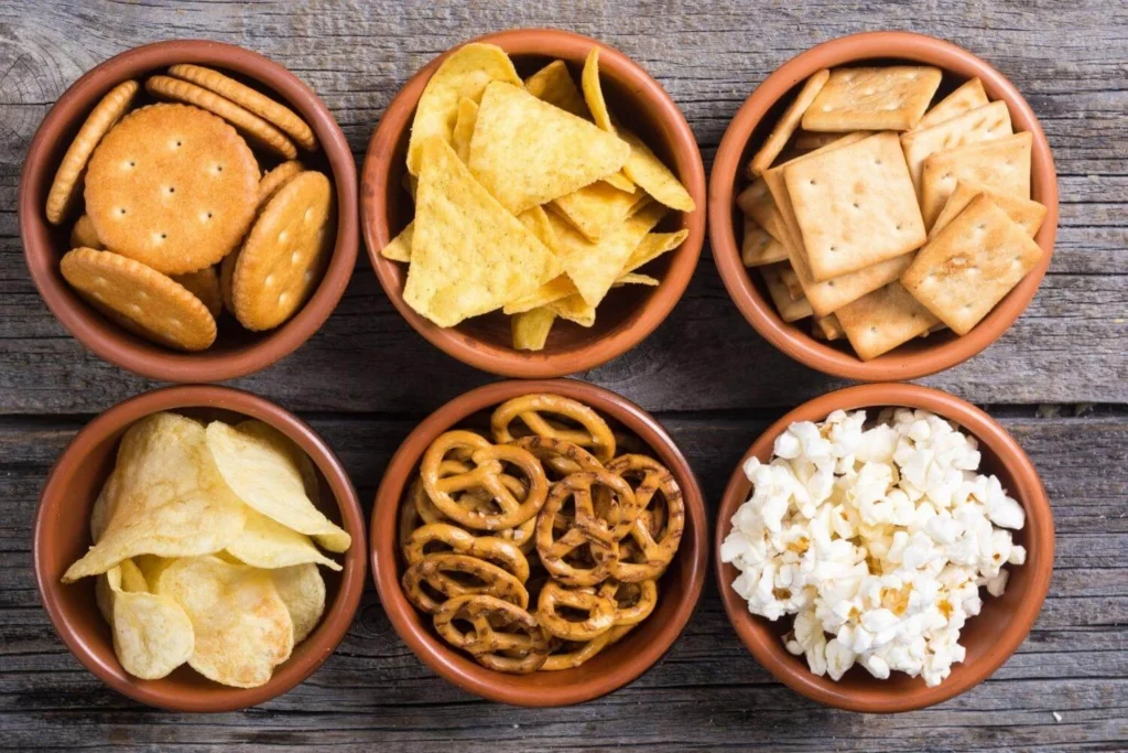 Assorted savoury snacks in small bowls including crackers, crisps, pretzels, tortilla chips, and popcorn, typical of British Christmas party food. 