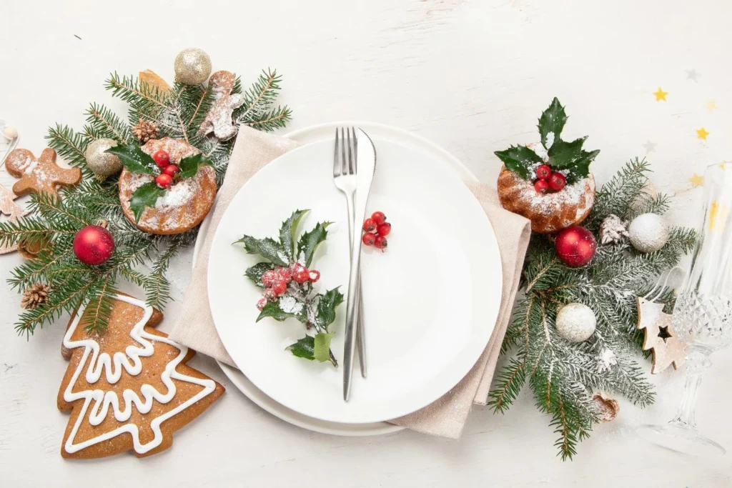 White plate with silver cutlery and holly garnish, surrounded by gingerbread cookies, mini cakes, and festive greenery, styled for a cozy Christmas solo meal.