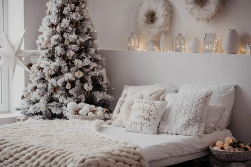 Minimalist bedroom with a flocked Christmas tree decorated in neutral tones, surrounded by fairy lights and soft textured pillows. A cozy knit blanket is draped across the bed for a wintery, calming vibe.