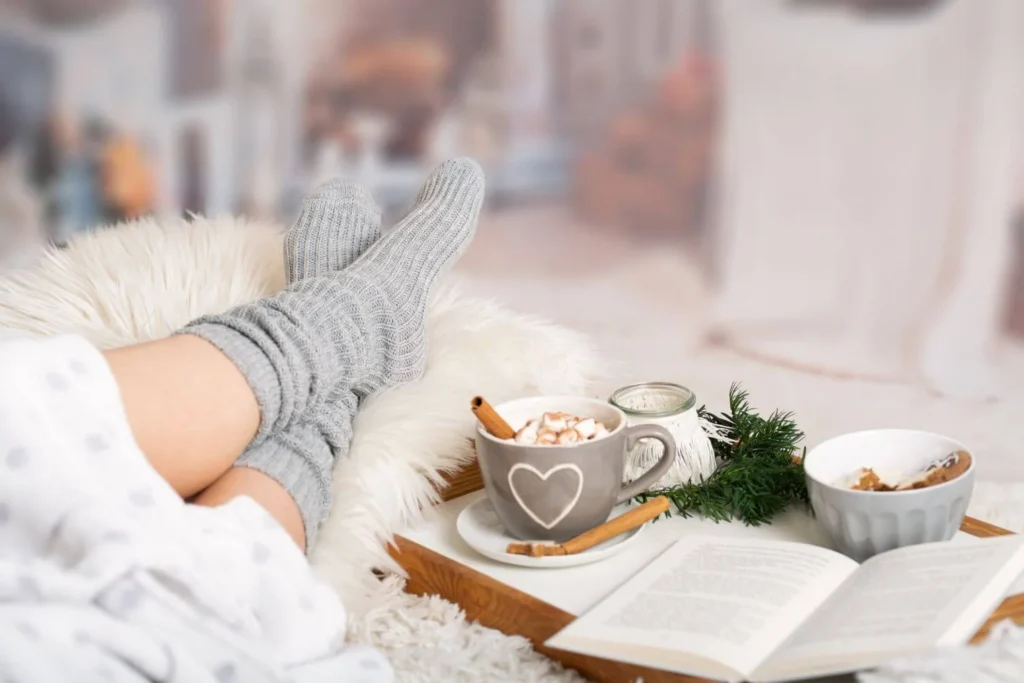 Person lounging in grey socks with a tray holding a mug of hot chocolate topped with marshmallows, an open book, and a bowl of snacks. The warm, festive setting includes fairy lights and a soft blanket.
