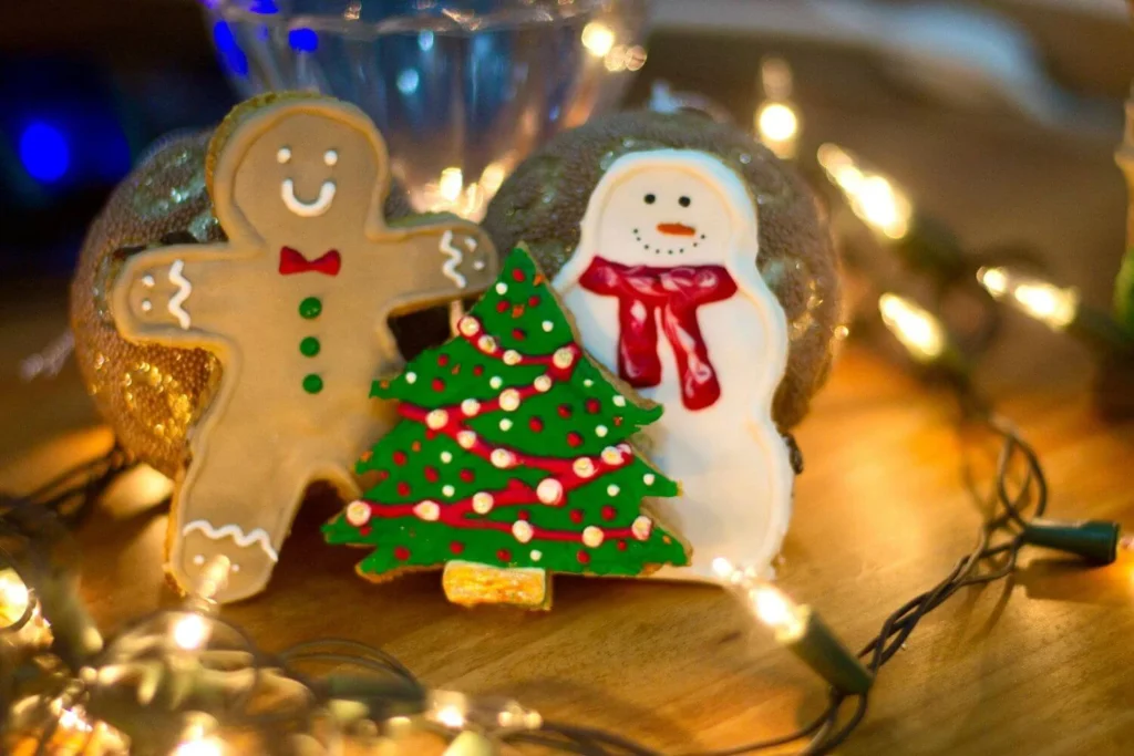 Close-up of Christmas cookies shaped like a gingerbread man, snowman, and tree with colorful icing, surrounded by string lights. A cozy holiday treat moment captured.