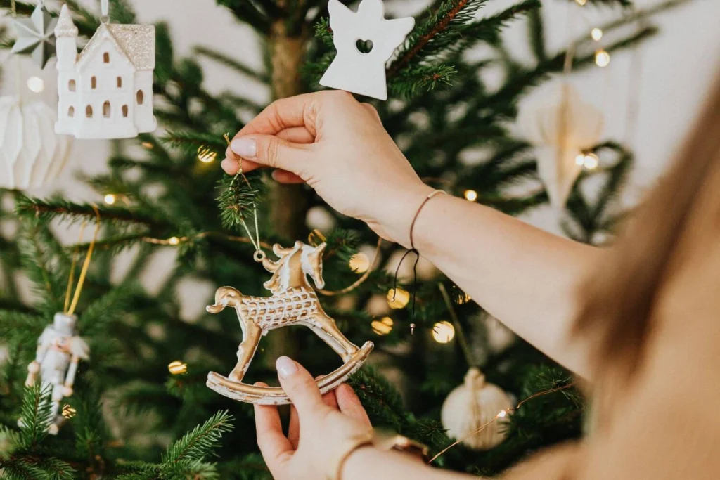 Close-up of hands hanging a vintage-style rocking horse ornament on a Christmas tree, with white and gold decorations in the background.