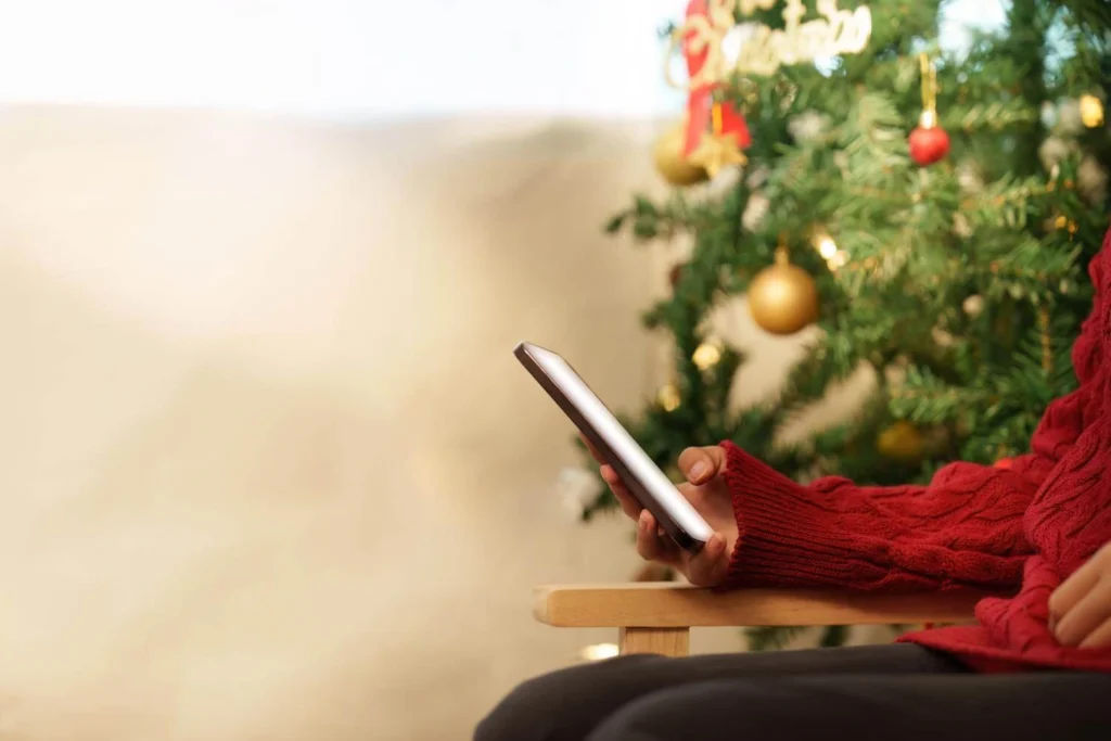 Person in a red sweater holding a phone while sitting next to a decorated Christmas tree, suggesting digital Christmas shopping.