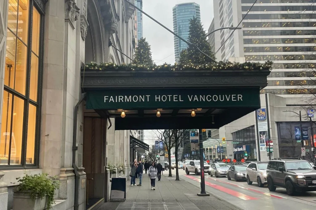 Street-level view of the Fairmont Hotel Vancouver entrance with a green awning, festive garland and Christmas trees on top, as pedestrians walk by and traffic lines the downtown street.