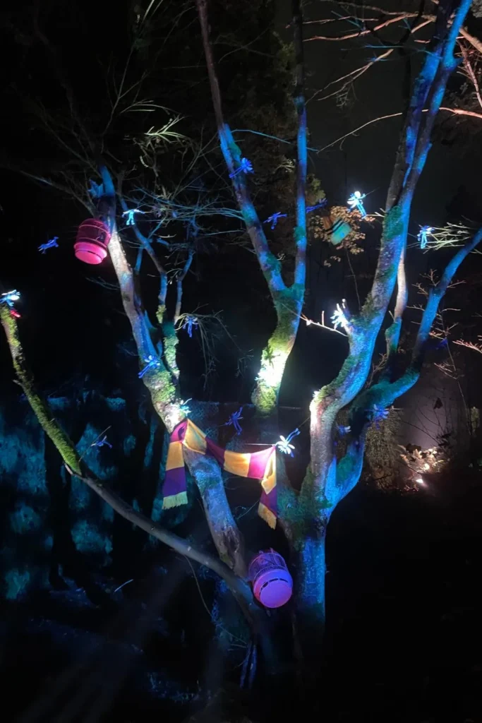 A tree decorated with glowing magical creatures, a hanging cage and a Gryffindor-style scarf illuminated in blue and purple light.