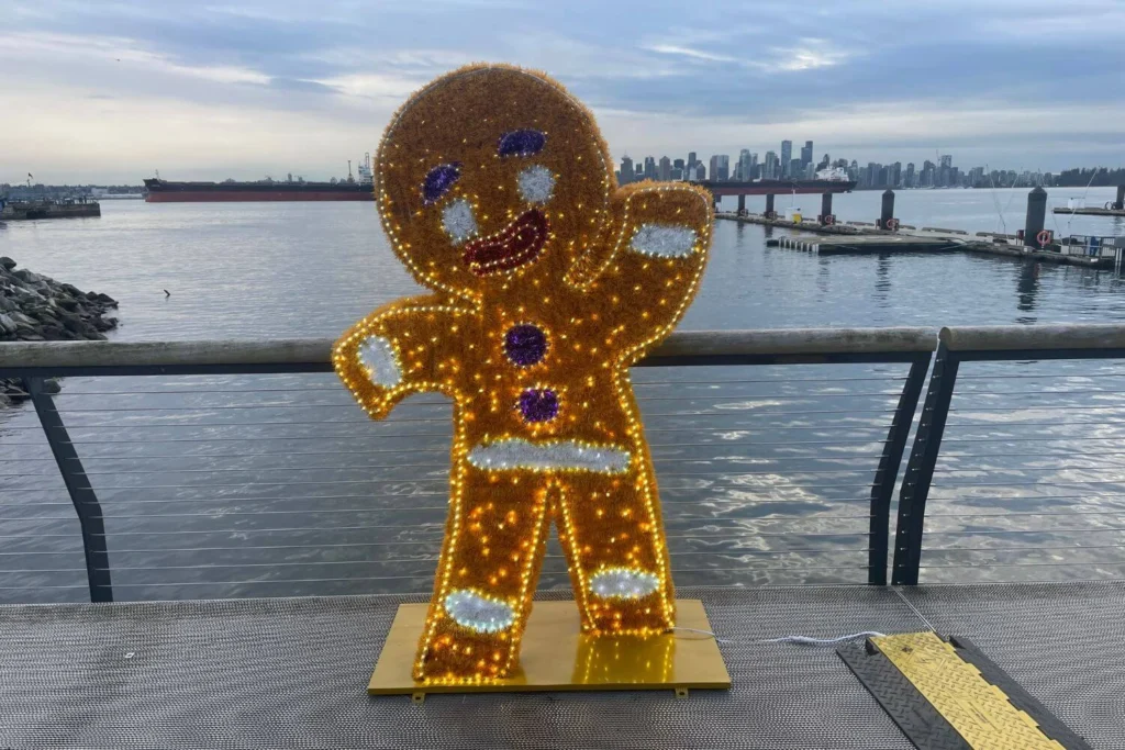 Cheerful gingerbread man sculpture made of Christmas lights on the North Vancouver waterfront, with the Vancouver skyline and Burrard Inlet in the background.