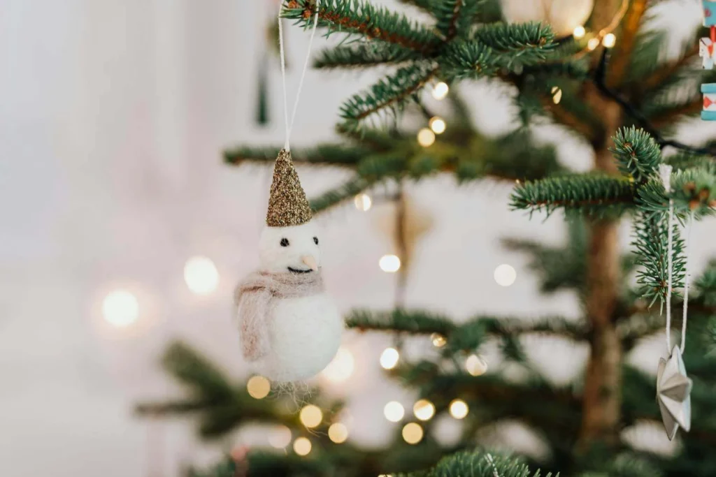 Close-up of a felted snowman ornament with a gold glitter hat hanging on a Christmas tree branch, surrounded by warm twinkle lights and minimalist decor.