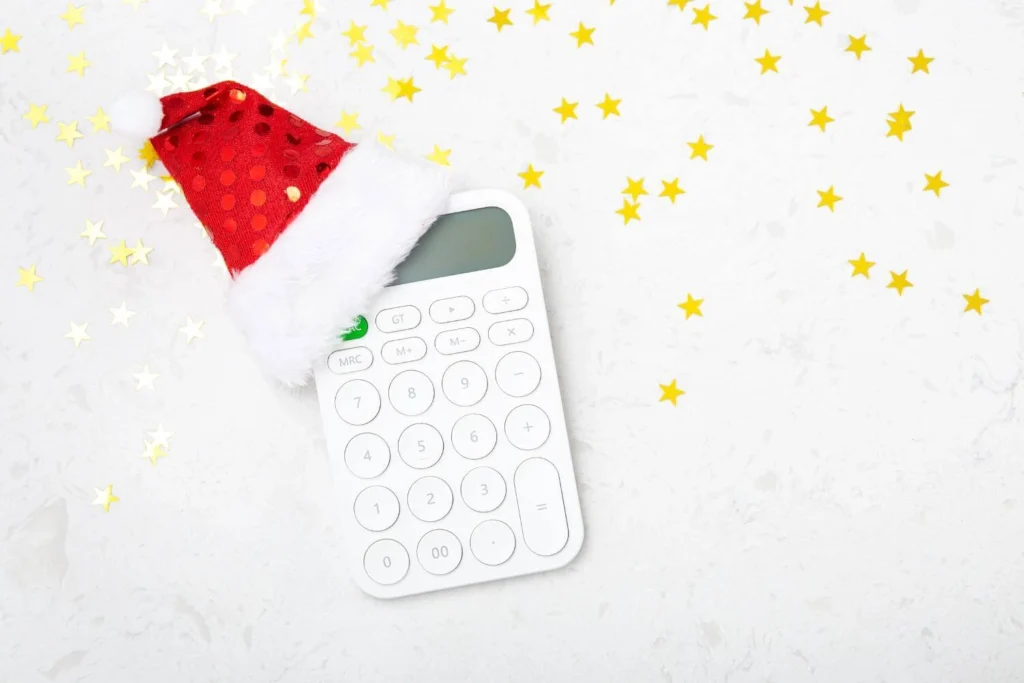 White calculator wearing a red Santa hat surrounded by gold star confetti on a marble background, representing end-of-year budgeting or Christmas expenses.