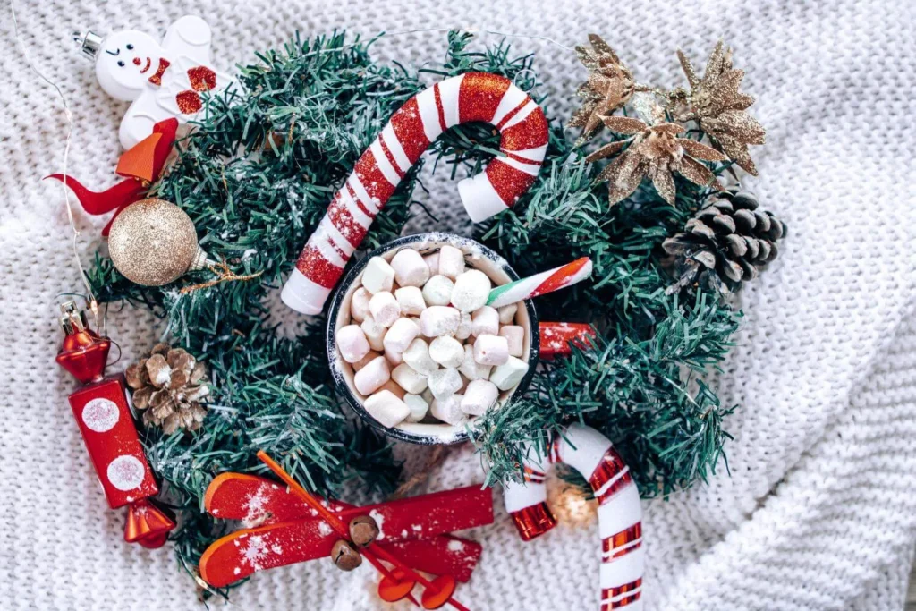 Flatlay of a cup of hot chocolate with mini marshmallows surrounded by Christmas garland, candy canes, ornaments, and cozy winter decor.