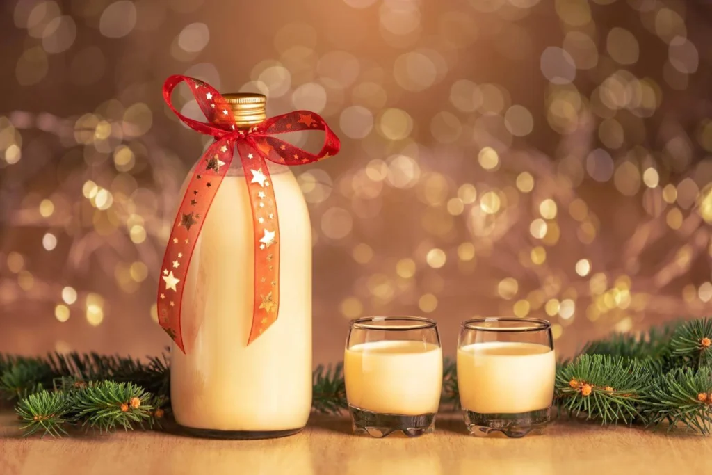 Bottle and glasses of British-style eggnog decorated with a festive ribbon and pine branches on a Christmas table.