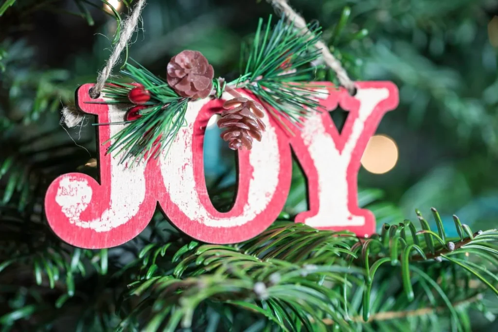 Red and white wooden "JOY" ornament hanging on a Christmas tree branch, adorned with pinecones and faux berries. Soft festive lights twinkle in the background.