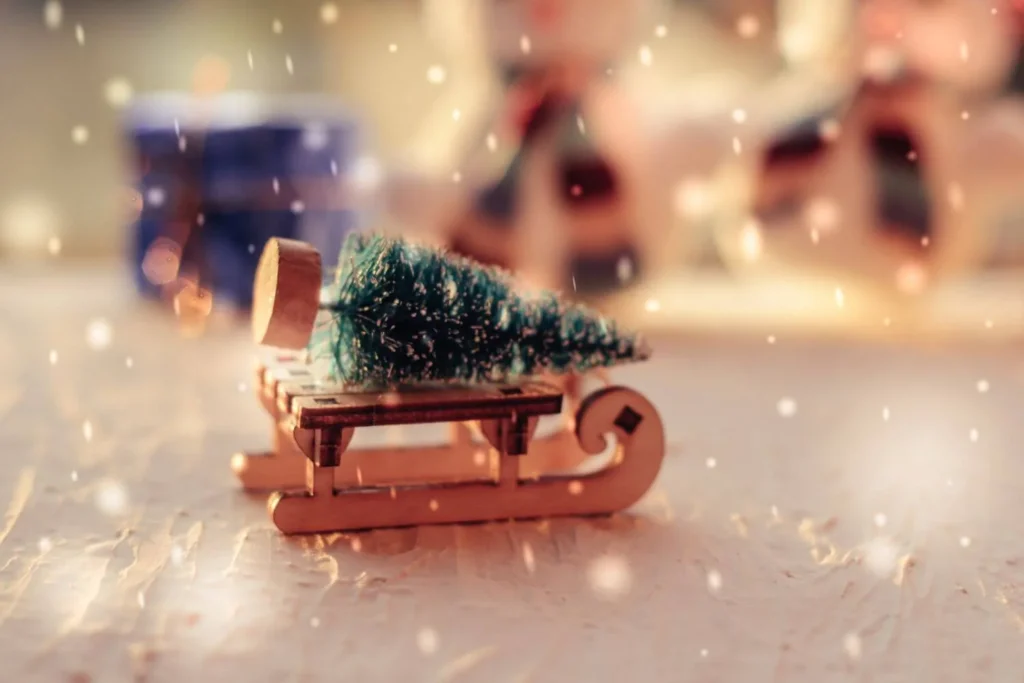 Tiny artificial Christmas tree tied to a miniature wooden sled, with soft snow-like lighting and blurred holiday figures in the background. A whimsical and nostalgic winter vignette.