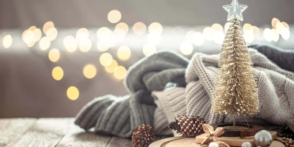 Small gold Christmas tree on a rustic wood surface with cozy folded knit blankets, pinecones, and bokeh holiday lights in the background, creating a minimalist festive atmosphere.