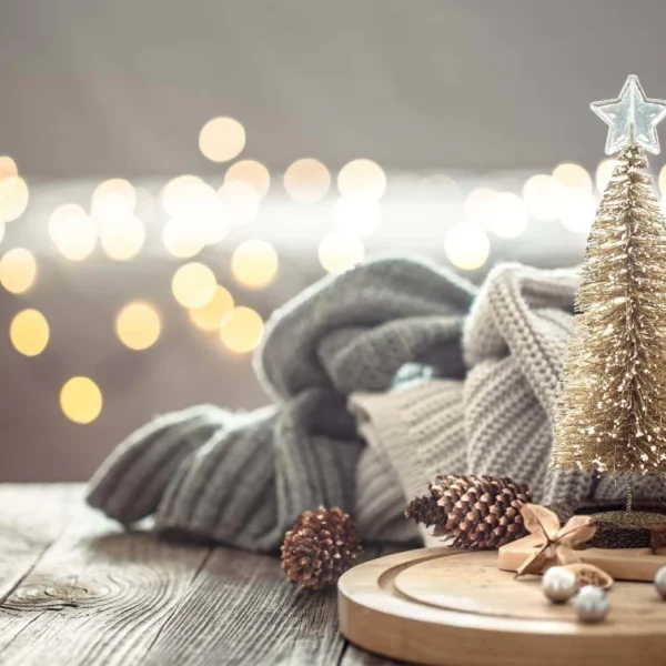 Small gold Christmas tree on a rustic wood surface with cozy folded knit blankets, pinecones, and bokeh holiday lights in the background, creating a minimalist festive atmosphere.