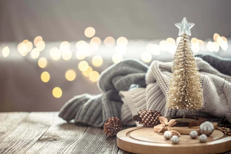 Small gold Christmas tree on a rustic wood surface with cozy folded knit blankets, pinecones, and bokeh holiday lights in the background, creating a minimalist festive atmosphere.