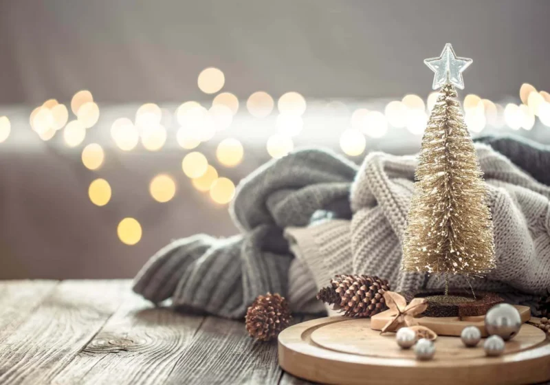 Small gold Christmas tree on a rustic wood surface with cozy folded knit blankets, pinecones, and bokeh holiday lights in the background, creating a minimalist festive atmosphere.