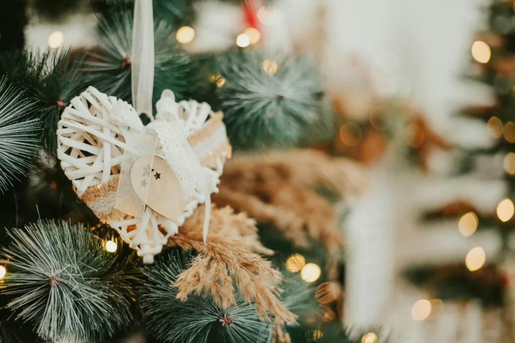 Close-up of a rustic heart-shaped Christmas ornament made of white wicker, burlap, and lace, hanging on a pine branch with warm fairy lights. The decor has a neutral, handmade charm.