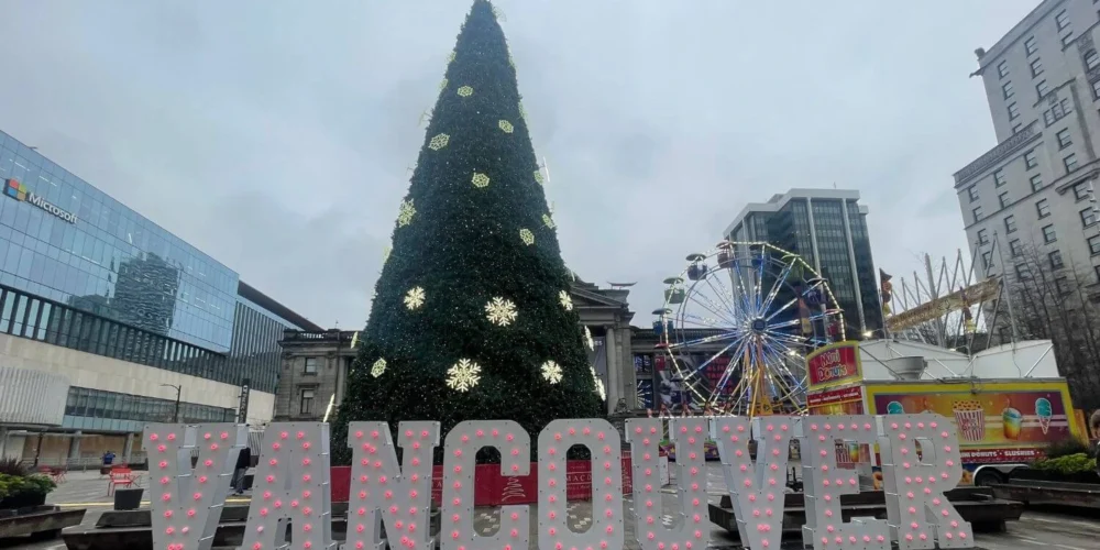 Giant Christmas tree decorated with glowing snowflakes stands behind large "VANCOUVER" light-up letters at Robson Square, with a Ferris wheel and colorful food truck nearby on a grey winter day.