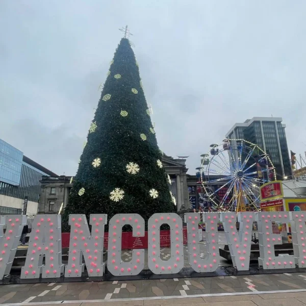Giant Christmas tree decorated with glowing snowflakes stands behind large "VANCOUVER" light-up letters at Robson Square, with a Ferris wheel and colorful food truck nearby on a grey winter day.