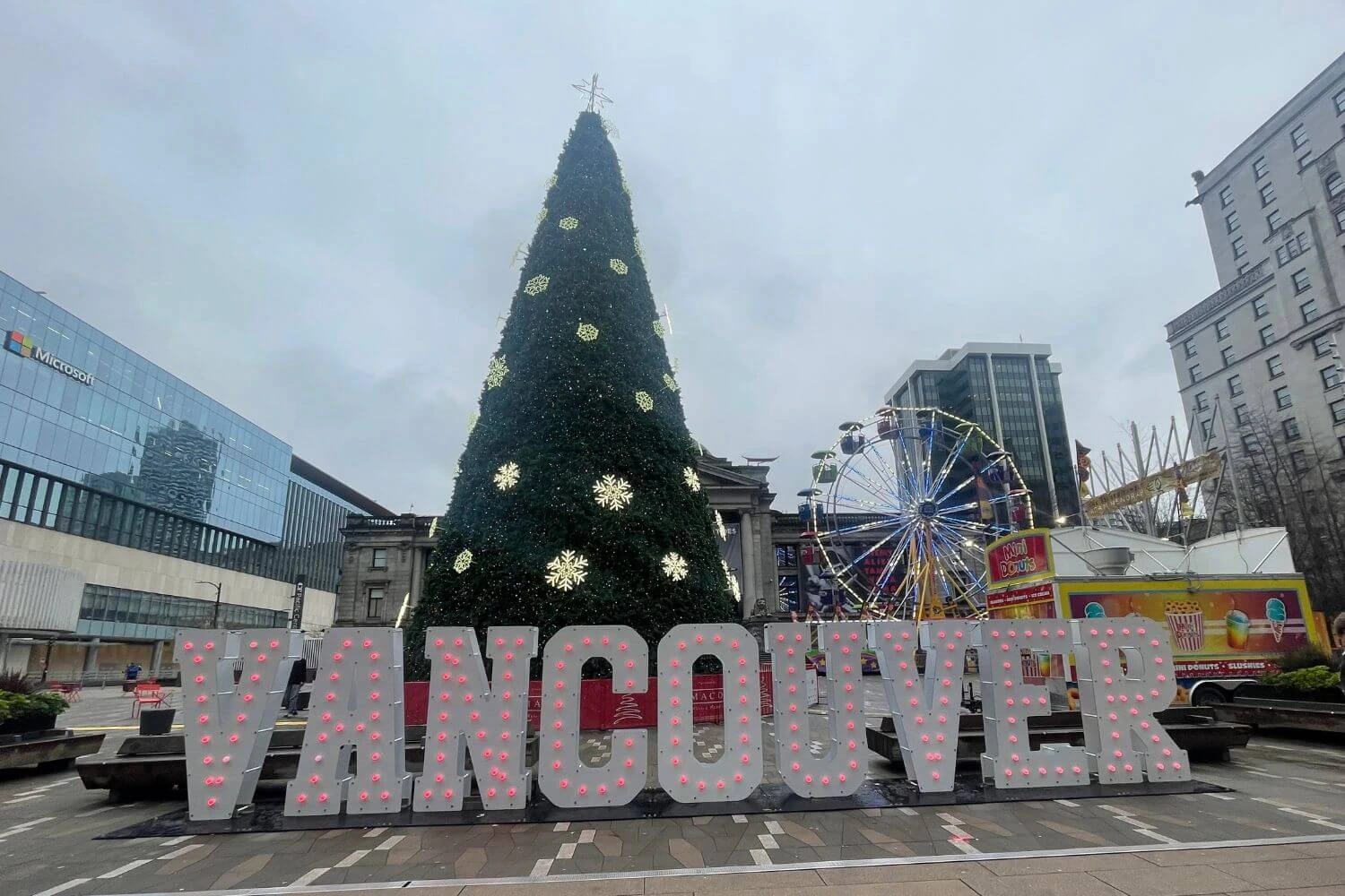 Giant Christmas tree decorated with glowing snowflakes stands behind large "VANCOUVER" light-up letters at Robson Square, with a Ferris wheel and colorful food truck nearby on a grey winter day.