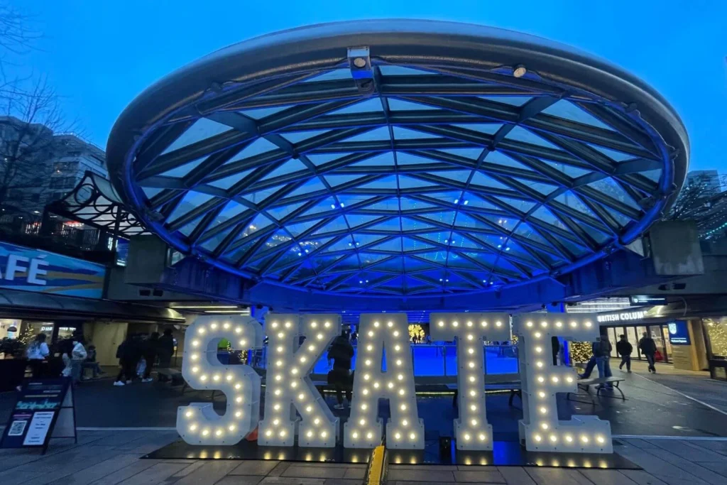 Large illuminated "SKATE" sign in front of the Robson Square ice skating rink in downtown Vancouver, with a glowing blue-lit glass dome overhead and people gathering around the area.