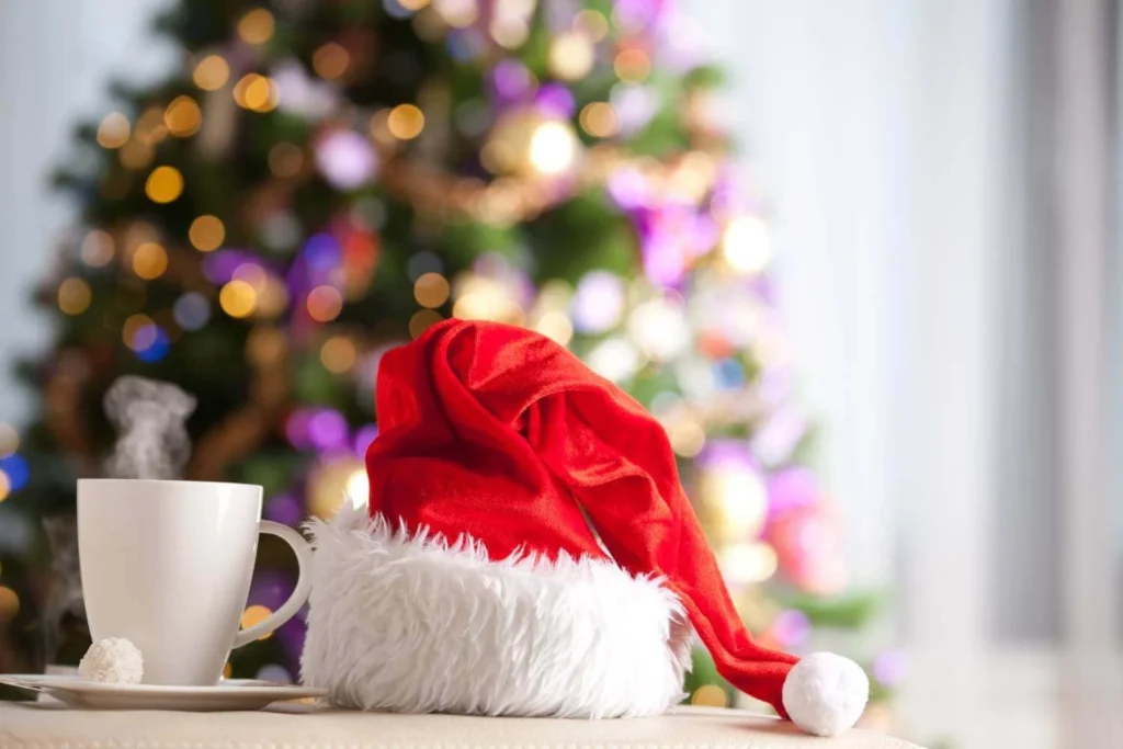 A fluffy Santa hat and a steaming mug of hot drink rest in front of a glowing Christmas tree with colorful lights, capturing a cozy holiday moment.