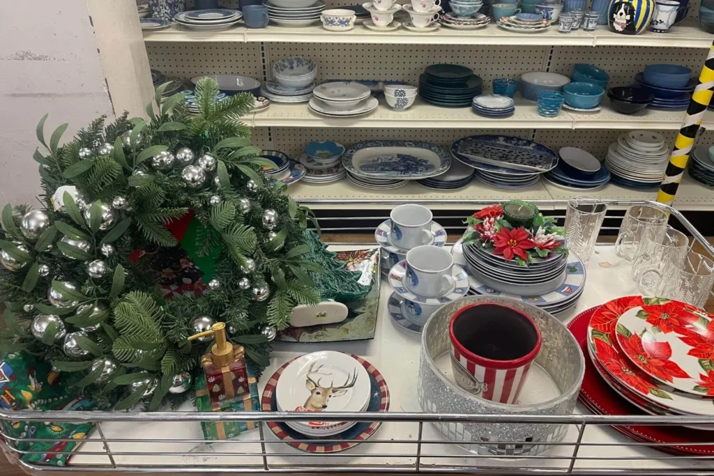 Secondhand Christmas dinnerware, glassware, and a silver-decorated wreath arranged on a table in front of thrift store shelves.