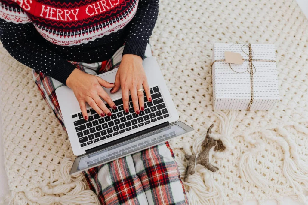 Person in festive pajamas and a Christmas sweater typing on a laptop while sitting on a cozy knitted blanket, with a wrapped gift and felt reindeer ornament nearby.