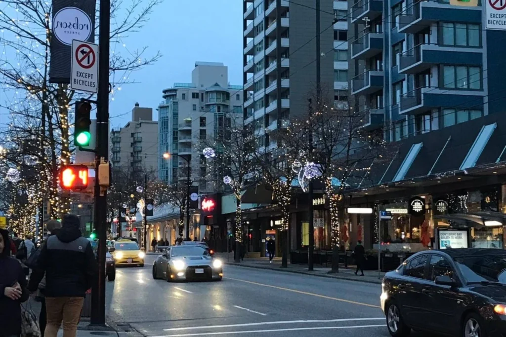 Robson Street in Vancouver at dusk with holiday lights on trees, pedestrians, and cars.