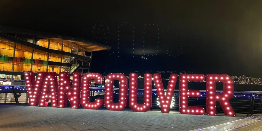 Large illuminated VANCOUVER sign glowing at the waterfront at night, with festive lights and the convention centre in the background during the holiday season.