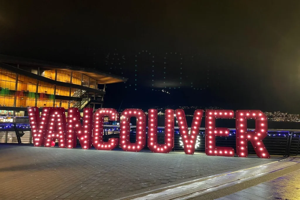Large illuminated VANCOUVER sign glowing at the waterfront at night, with festive lights and the convention centre in the background during the holiday season.
