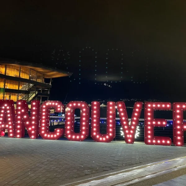 Large illuminated VANCOUVER sign glowing at the waterfront at night, with festive lights and the convention centre in the background during the holiday season.