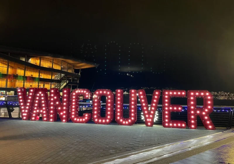 Large illuminated VANCOUVER sign glowing at the waterfront at night, with festive lights and the convention centre in the background during the holiday season.