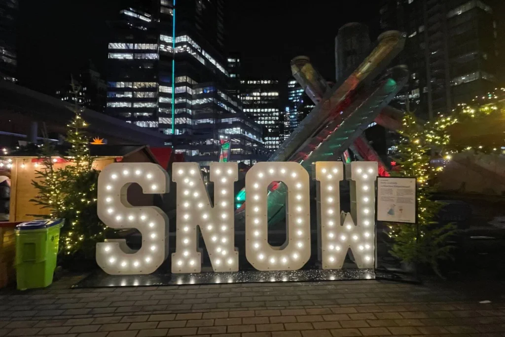 Bright SNOW sign lit up at Jack Poole Plaza in downtown Vancouver, with Christmas market stalls, trees, and city buildings in the background.