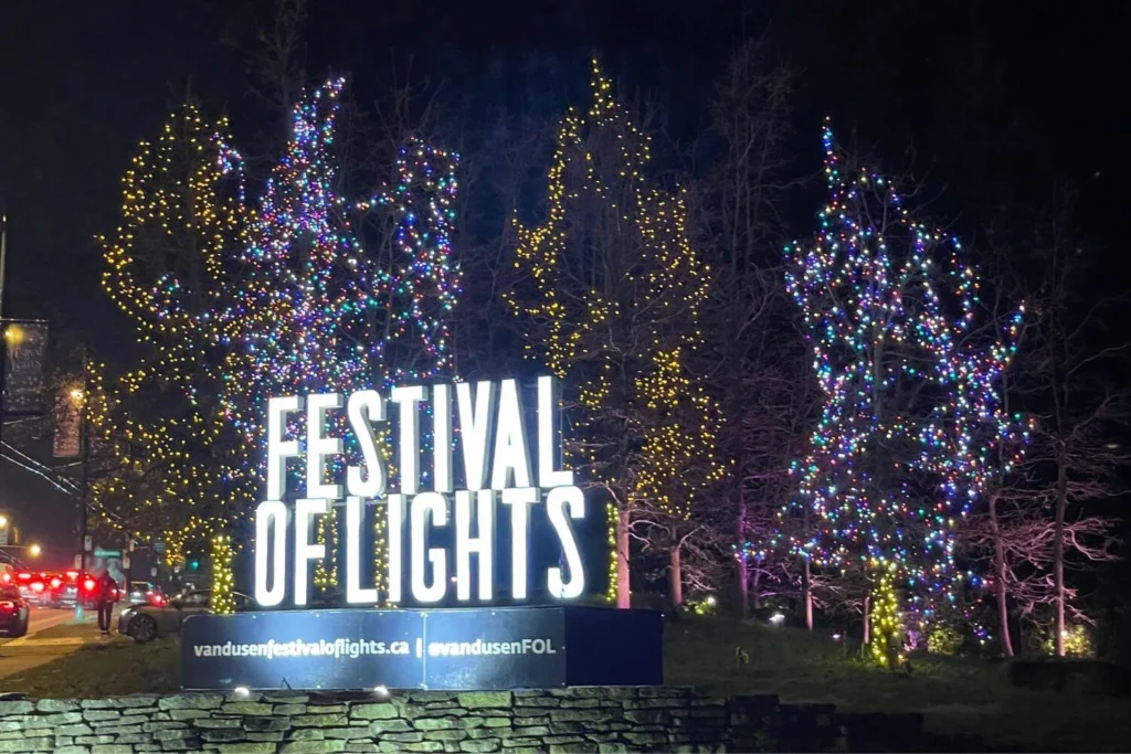 Festival of Lights entrance sign at VanDusen Botanical Garden in Vancouver, with colorful Christmas lights wrapped around bare trees at night.