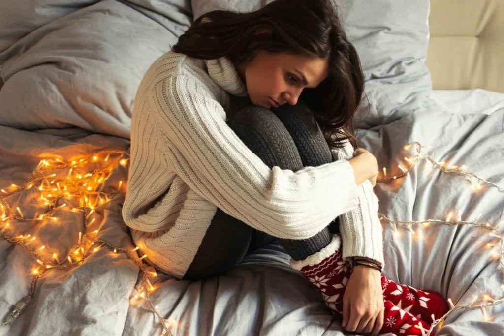 Young woman sitting curled up on a bed in cozy clothes surrounded by Christmas lights, looking pensive and lonely during the holidays.