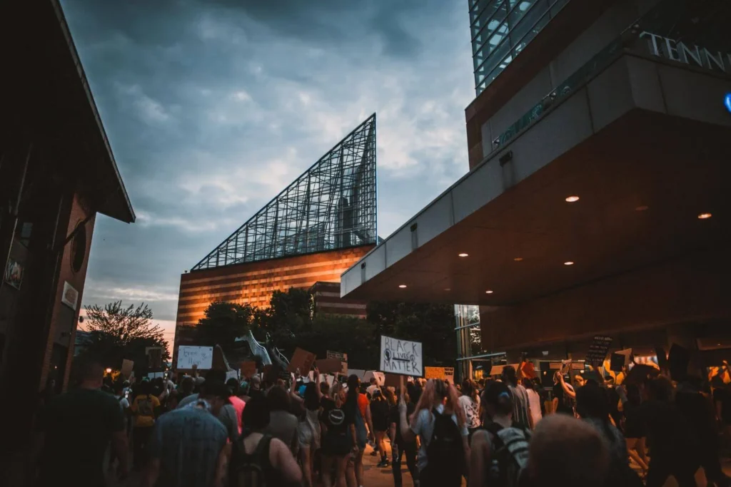Large crowd gathered at a public protest in downtown Chattanooga, Tennessee, with signs raised outside modern city buildings.