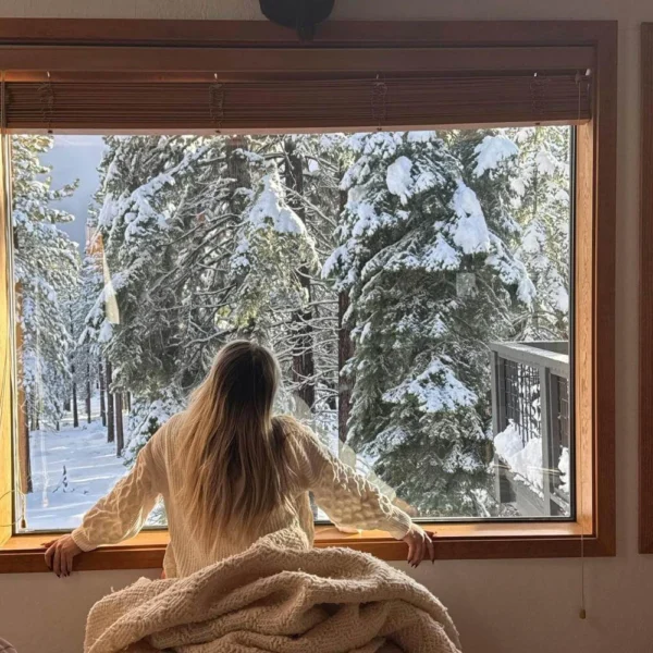 Solo traveler spending winter alone at home, looking out at snowy trees and reflecting quietly.