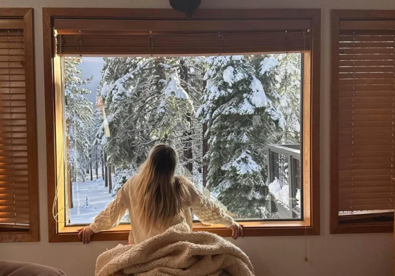 Solo traveler spending winter alone at home, looking out at snowy trees and reflecting quietly.
