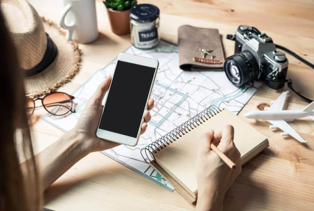 Solo traveler planning a trip with a phone, notebook, map, and travel essentials laid out on a desk.