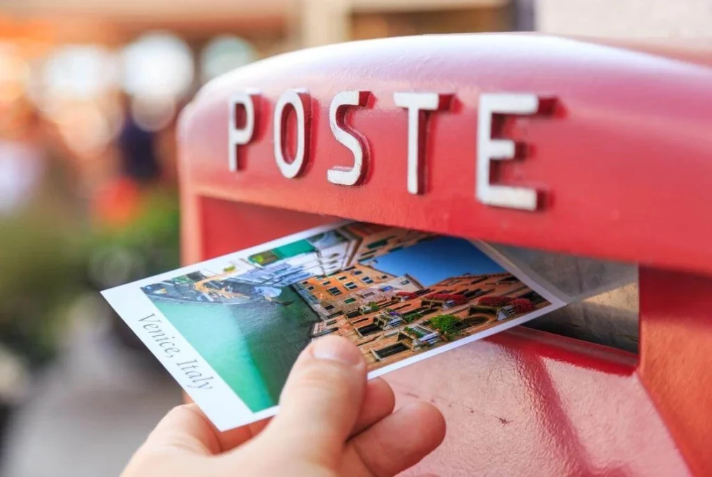 Hand placing a postcard labeled "Venice, Italy" into a red post box. The close up photo highlights the connection between travel experiences and the stories people choose to share through postcards.